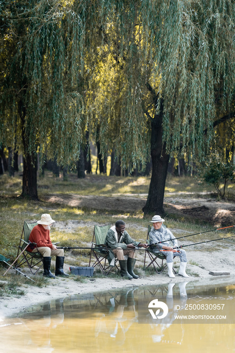 Cheerful multicultural men in rubber boots fishing on chairs near lake in park