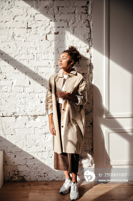 Indoor shot of african woman in trench coat and white sneakers. Sensual black girl enoying sunshine with closed eyes.