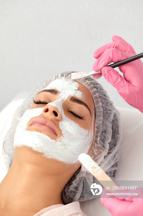 Satisfied young Caucasian beautiful woman uses services of professional beautician in beauty salon or spa. Unrecognizable beautician applied white mask to half of client’s face with brush. Top view.