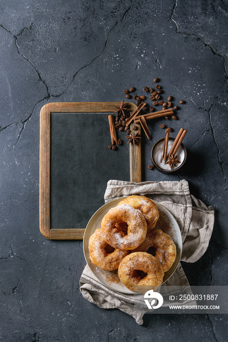 Plate of homemade donuts with sugar and cinnamon powder on vintage chalkboard served with spices, coffee beans and textile napkin over dark texture background. Top view with space for text