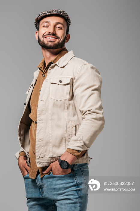 bearded smiling man in autumn jacket and tweed cap posing with hands in pockets, isolated on grey