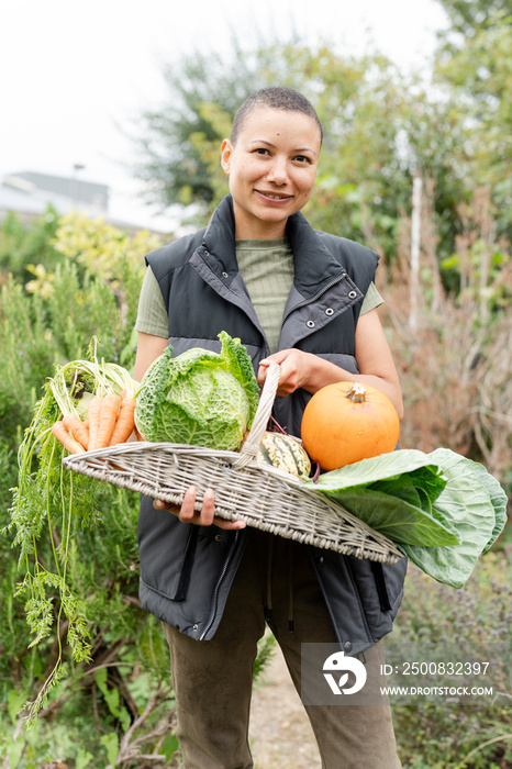 Portrait of smiling woman holding basket with fresh vegetables in urban garden