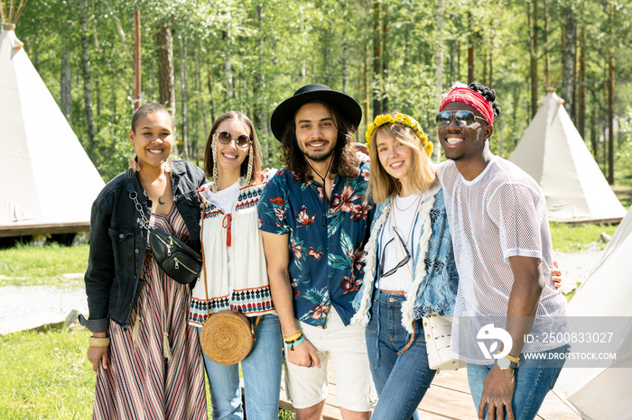 Portrait of cheerful young multi-ethnic friends in hippie outfits embracing each other at festival in forest campsite