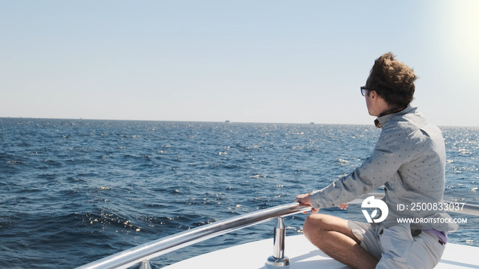 Man on yacht looks in distance, sunny morning. White sailing boat on blue sea waves, handsome man in light clothes, sunglasses sitting on bow. Luxury voyage summer travel adventure. Nautical lifestyle