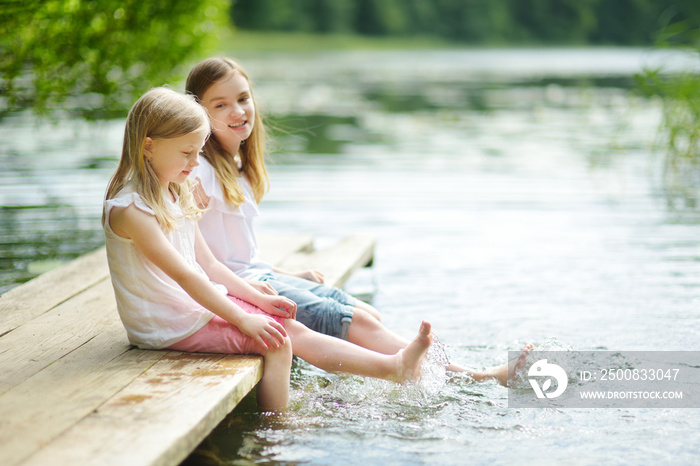 Two cute little girls sitting on a wooden platform by the river or lake dipping their feet in the water on warm summer day