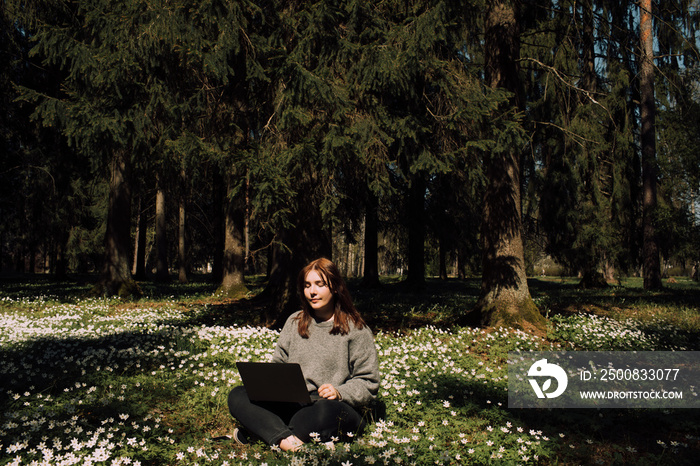 Woman with a laptop in the park. Young female sitting in grass with flowers work remotely in nature. Work everywhere travel blogger outdoors. Businesswoman sitting in forest Remote working concept