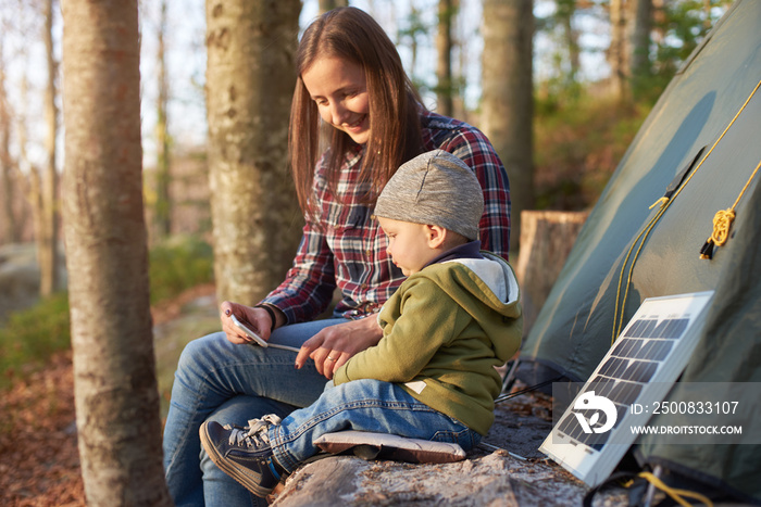 Mother with son on camping vacation looking at a mobile phone while enjoying the fresh air. Family sit on a log near the tent and tourist solar panel in the forest on a sunny day.