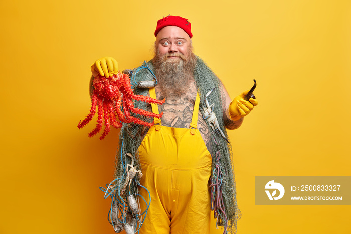 Expressive cheerful bearded sailor man holds big octopus, carries smoking pipe, fishing net, enjoys summer vacation, dressed in overalls, poses against yellow background. Overweight fisherman