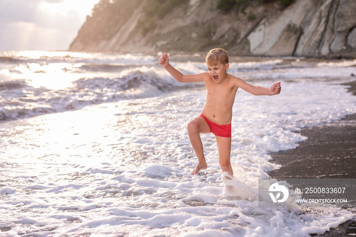 Blonde boy running and jumping on the beach on blue sea shore in summer vacation at the day time. Blue ocean with white big wawes on the background