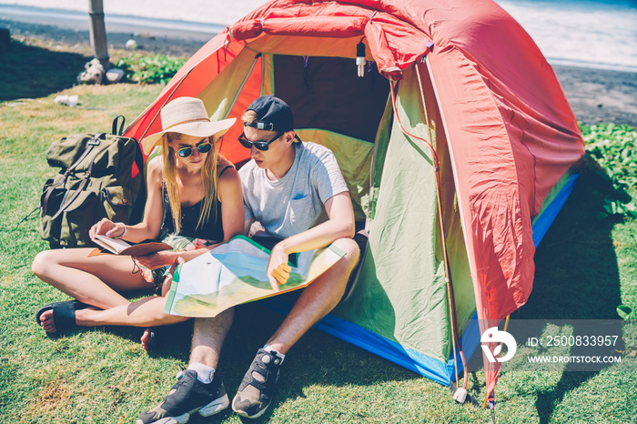 Concentrated male and female travelers discussing ideas for route using notes and map sitting in tent,hipster couple camping on green grass near beach reading guide for choosing direction for journey