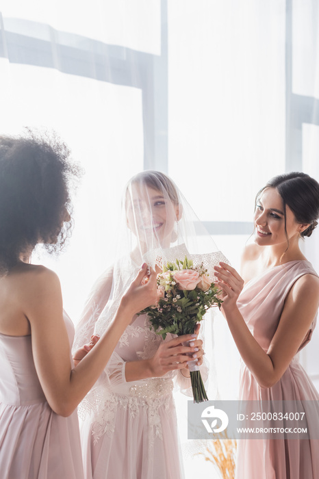 multicultural bridesmaids touching veil of smiling bride holding wedding bouquet.