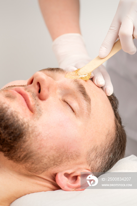 Beautician applying wax paste between eyebrows during the procedure of waxing in the beauty salon