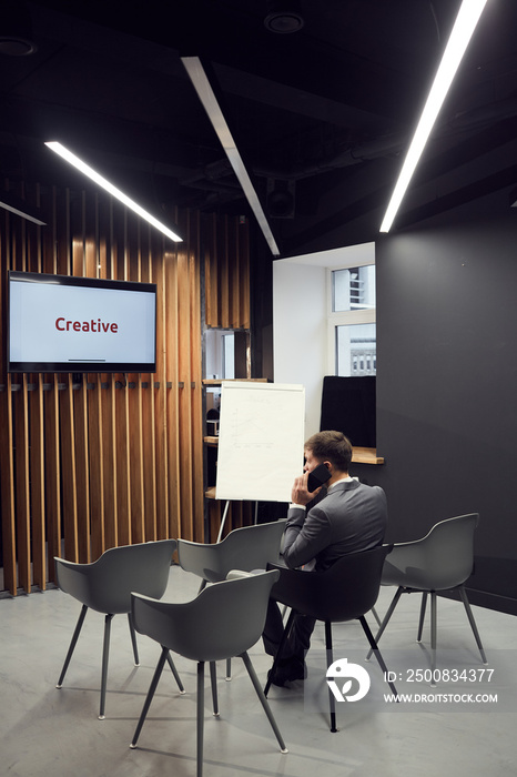 Rear view of busy young businessman in suit sitting on chair in conference room and calling by cell phone, Creative inscription on plasma screen