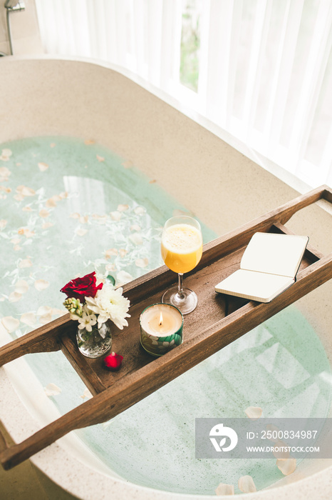 Bright Bathroom at Home. Bath with Flower Petals and Salt. Tray over the Bath with Orange Juice, Notepad and Candles.