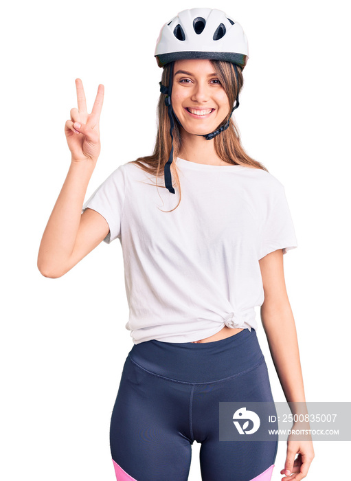 Young beautiful girl wearing bike helmet smiling with happy face winking at the camera doing victory sign. number two.