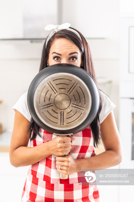 Young girl covering her mouth and nose with a pan, with eyes wide open, surprised face expression, standing in the kitchen