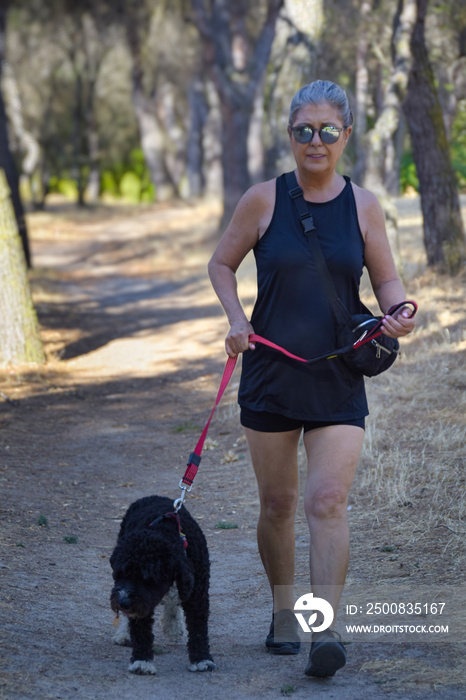 Mujer madura de pelo gris y con gafas de sol paseando por un parque con un perro negro.