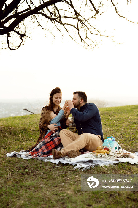 a young pregnant woman with a handsome husband and a little girl are on a picnic at sunset. husband and wife laugh and drink juice. family time