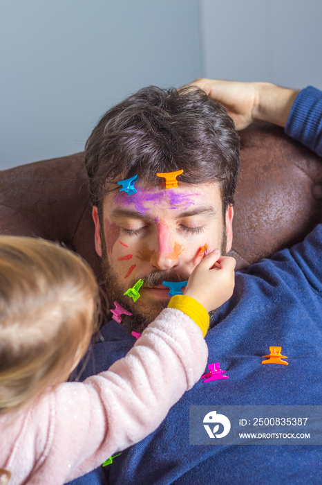 Father’s Day celebration. Young man sleeping on the sofa while his little daughter makeup her face with colorful watercolors and hair clips