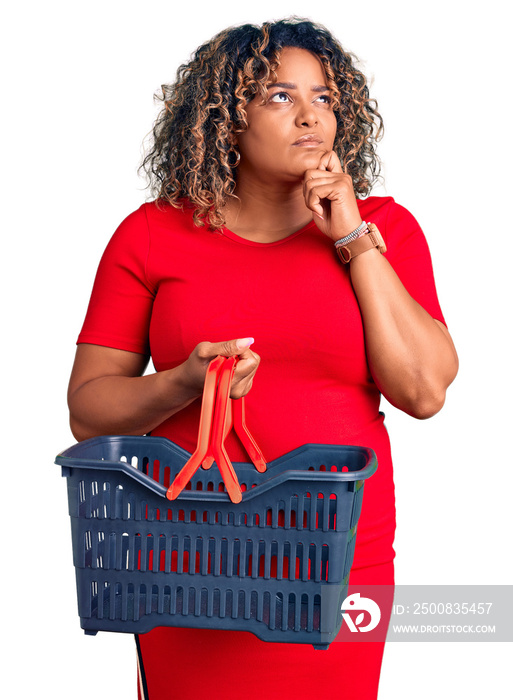 Young african american plus size woman holding supermarket shopping basket serious face thinking about question with hand on chin, thoughtful about confusing idea