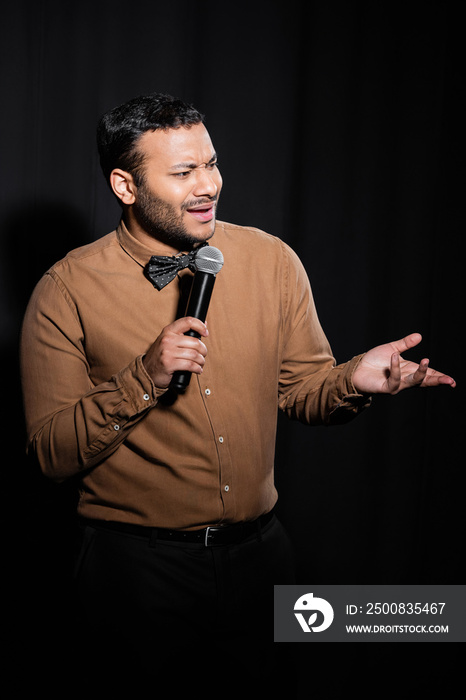 displeased indian stand up comedian in shirt and bow tie holding microphone during monologue on black.