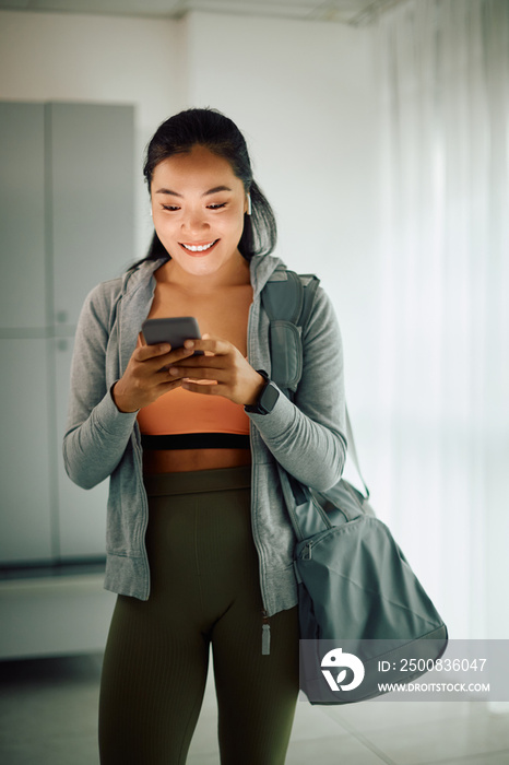 Happy Asian sportswoman uses mobile phone while text messaging in locker room at gym.