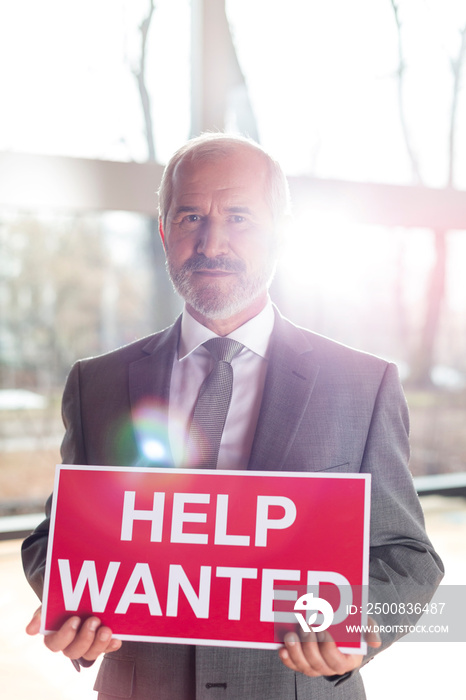 Portrait of confident senior businessman holding help wanted sign while standing at office