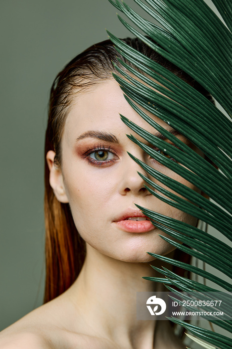 a close beauty portrait of a beautiful woman standing holding a tropical palm leaf in her hand, bringing it to her face, looking into the camera. Vertical photo without retouching of problem skin