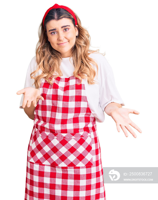 Young caucasian woman wearing apron clueless and confused expression with arms and hands raised. doubt concept.