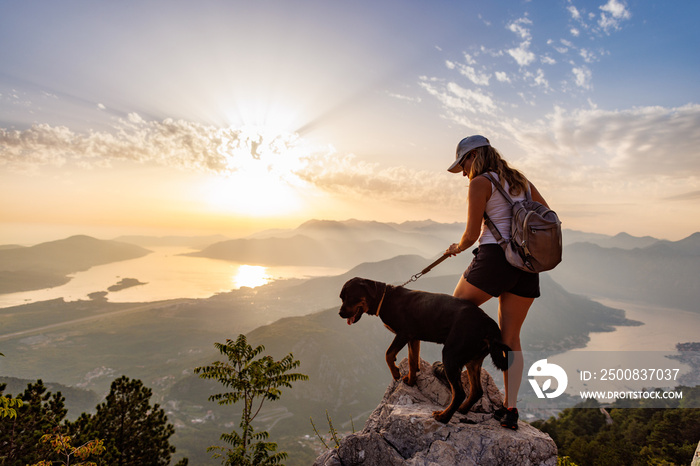 A sporty girl with a backpack stands on the edge of a mountain with a Rottweiler dog