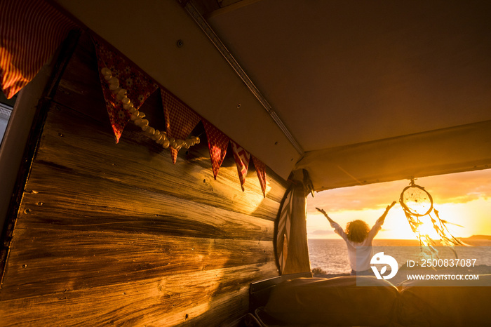 Rear view of woman with arms outstretched admiring scenic seascape during sunset next to dreamcatcher hanging from the camper van. Woman with curly hair enjoying her vacation at beach by dreamcatcher