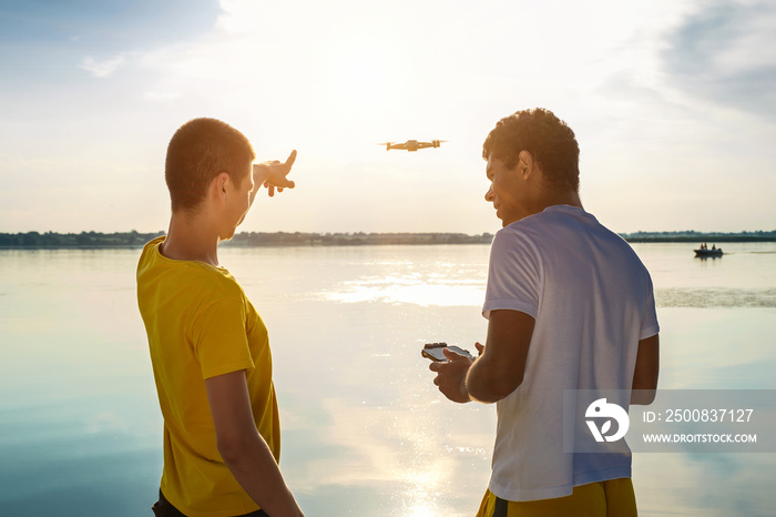 Young smiling African American man and his caucasian friend operating drone with controller and standing near the river at sunset