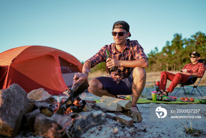 Happy young friends enjoy a sunny day at the nature. They’re toasting with beer bottles by the bonfire near tent.