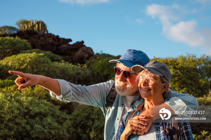 Cheerful senior caucasian couple hugging in mountain trekking enjoying nature and freedom. Smiling old retirees in hat and casual clothes among green bushes and blue sky