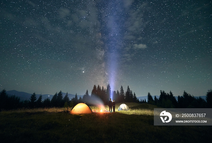 Night starry sky over grassy hill with illuminated camp tents and couple near campfire. Travelers standing near bonfire under sky with stars. Concept of travelling, night camping and relationship.