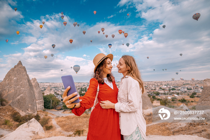 Two friends share their adventure in Cappadocia, Turkey on social networks with their smartphone selfies surrounded by breathtaking rock formations and hot air balloons.