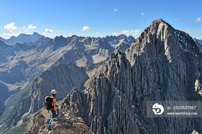 Female climber standing in wild rocky landscape with steep mountains under blue sky at a beautiful day in autumn. Dremelspitze, Lechtal Alps, Tyrol, Austria.