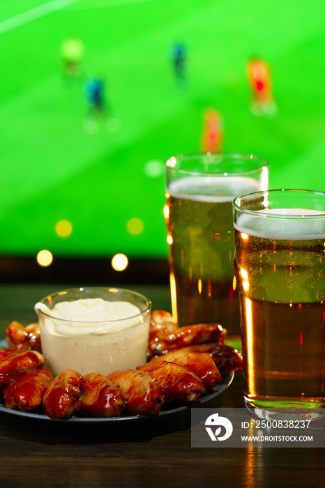 Hot barbecue chicken wings with 2 beer glasses on a dark wooden table served with honey mustard sauce. Football on a background, high resolution