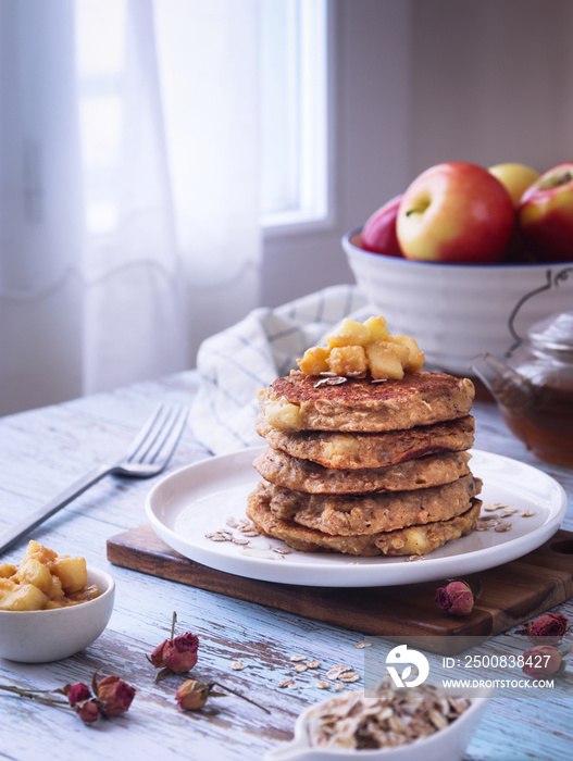 A plate of healthy rolled oat and apple pancakes topped with cooked diced apple pieces with a bowl of tis fruit and fork with teapot in background near the window with light.