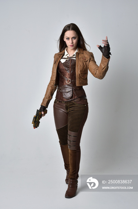 full length portrait of brunette  girl wearing brown leather steampunk outfit. standing pose, holding a gun. on grey studio background.