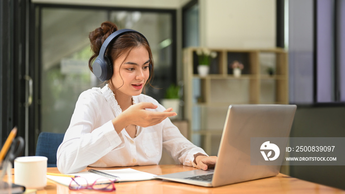 Young female employee wearing headphone and communicating by video conference on laptop computer