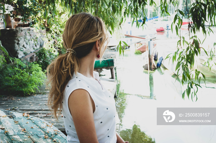 side view of woman in white blouse with ponytail looking at boats and sitting in front of tree branches near river