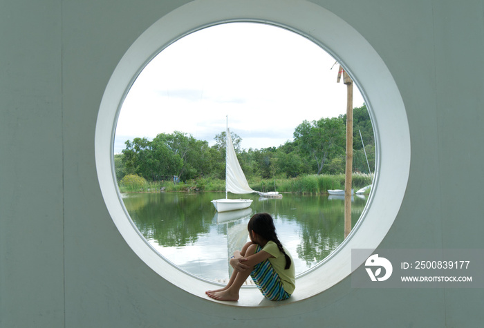 Asian girl sits looking at the view of a sailing boat from a circular aperture