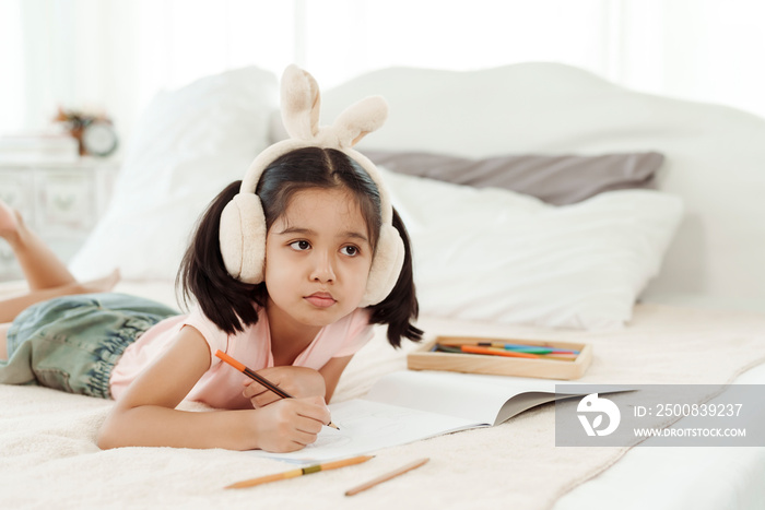 Cute little girl having fun in cute kindergarten age. Use colored pencils to draw a picture on the paper. Happily put the rabbit headphones on the bed. Concept family and happy learning