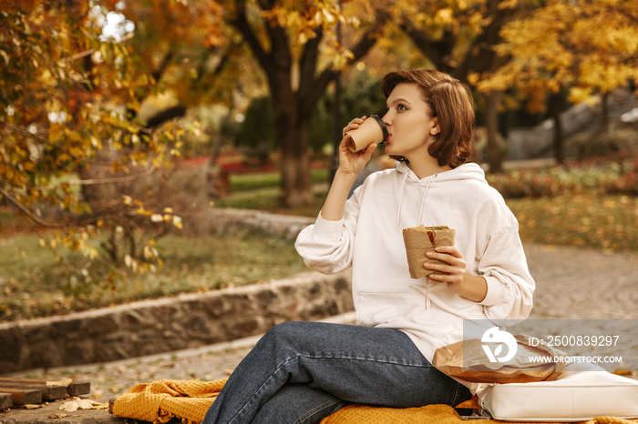 Cute fair-skinned young lady is drinking coffee, eating bun while sitting on park bench, in colorful autumn. Student has breakfast for breaks between classes. Free time concept