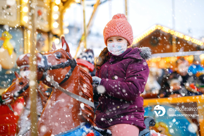 little kid girl with medical mask on face riding on merry go round carousel horse at Christmas funfair or market. masks as protection against corona virus. Covid pandemic time in Europe and the world