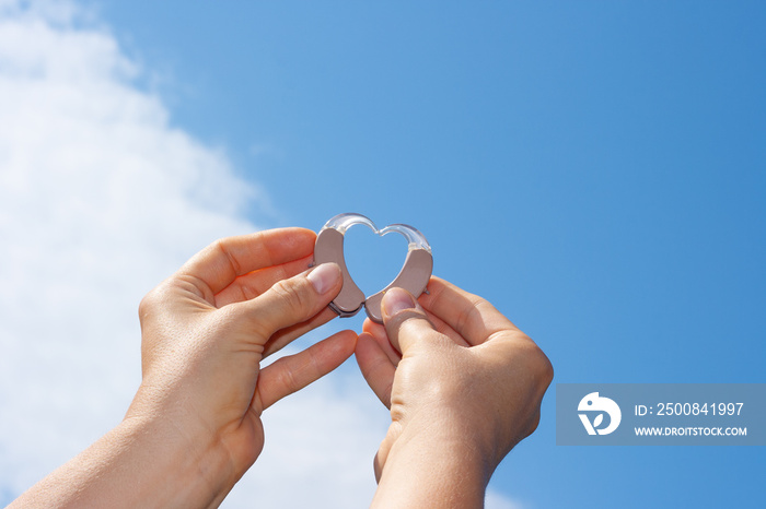 Woman’s hands showing heart with hearing aids in front of natural blue sky