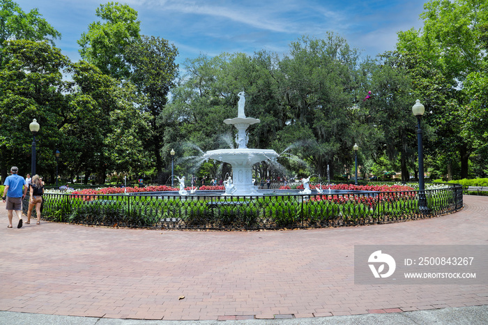 a circular white water fountain in the park  surrounded by colorful flowers and lush green trees and plants with people walking and sitting around the fountain at Forsyth Park in Savannah, Georgia