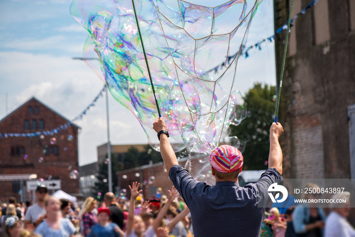 A freelance clown blowing hundreds of tiny, small and big bubbles at outdoor festival in city center. Concept of entertainment, birthdays. Kids having fun. Shower of bubbles flying in the happy crowd