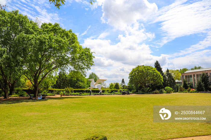 a white statue of a horse with wings in the garden surrounded by lush green trees, grass and plants with workers gardening in the plants and blue sky with powerful clouds at Atlanta Botanical Garden